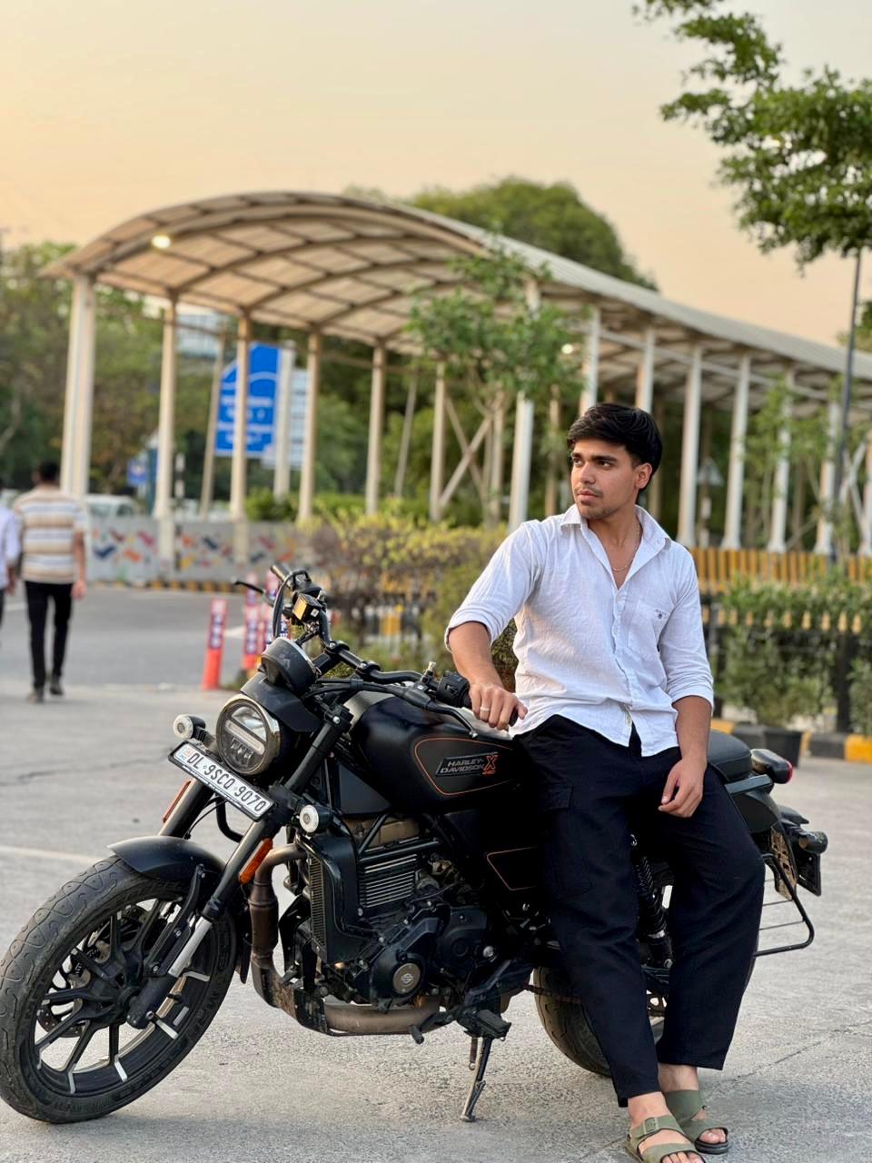 Stylish young man sitting on a black motorcycle outdoors during sunset, wearing a white shirt and black pants, posing confidently on a city road with protien snacks